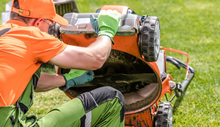 A man in an orange shirt and green gloves is fixing a lawn mower. He is troubleshooting lawn mower issues.